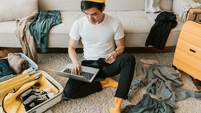 Young man casting vote on laptop while sitting on floor surrounded by packed suitcase in an ambiguous situation