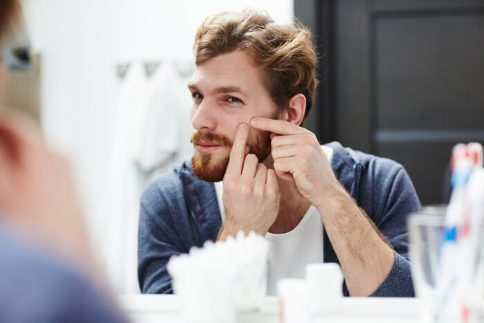 Man with red hair and beard squeezing a pimple in front of a bathroom mirror in an embarrassing moment.