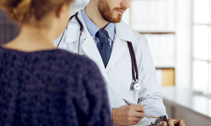 Doctor with stethoscope writing notes while a patient recounts embarrassing moments in front of doctors during a consultation.