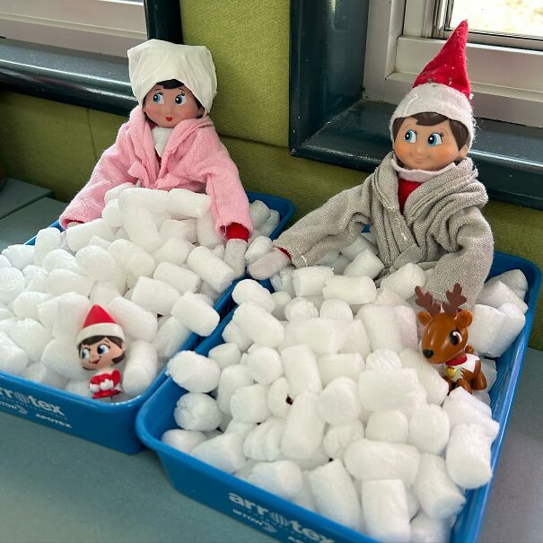 Two Elf on the Shelf dolls in bathrobes sitting in bins filled with packing peanuts for a fun elf on the shelf idea.