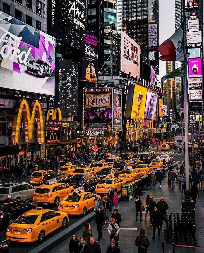 Yellow taxis and crowds on a busy New York City street lined with illuminated billboards and vibrant signage.