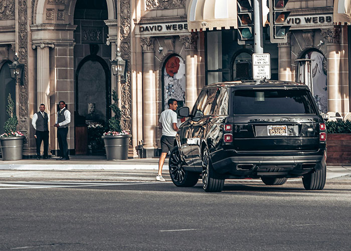 Luxury industry scene with a black SUV and doormen outside an upscale building highlighting rip-offs the rich pay for.