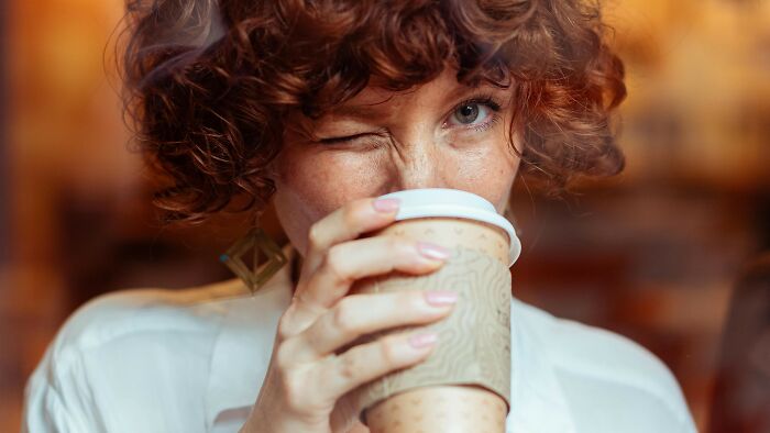 Young woman with curly hair winking while holding a takeaway coffee cup, engaging in ambiguous social situations discussion.