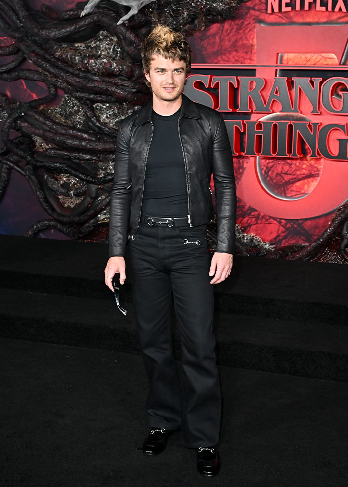 Male celebrity wearing an all-black outfit with leather jacket and flared pants at a 2025 fashion event.