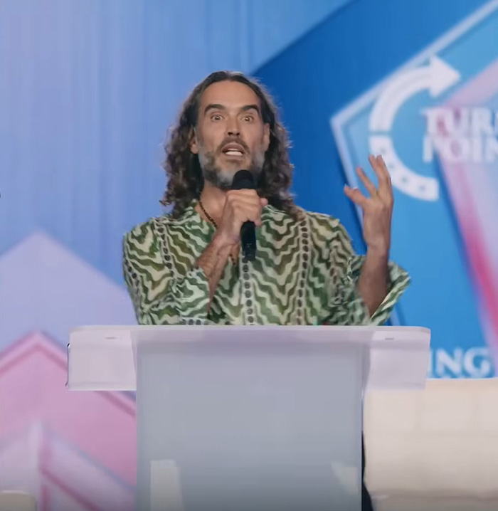 Russell Brand speaking passionately at a podium, reacting bitterly to Katy Perry and Justin Trudeau news.