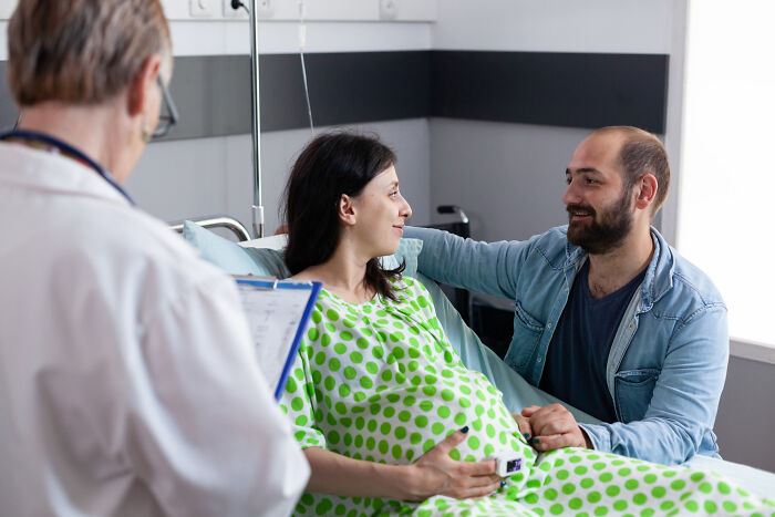 Man supporting pregnant partner in hospital room while doctor reviews notes, illustrating men missed delivery babies concept.