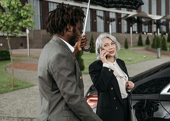 Woman talking on phone near car while man holds umbrella, illustrating people who work in luxury industries and rip-offs.