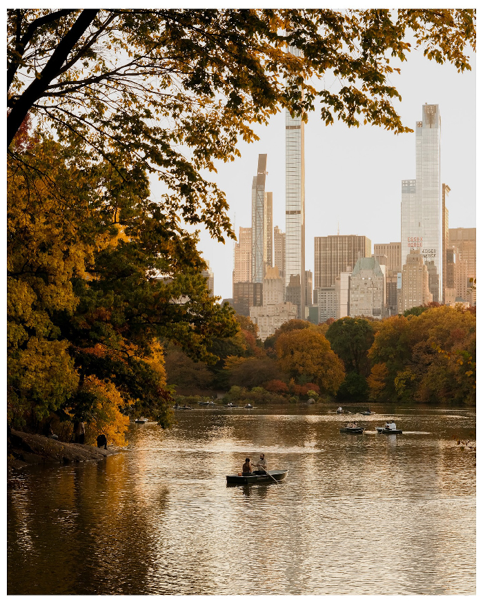 Autumn view of Central Park lake with rowboats and NYC skyscrapers in the background on a calm day.