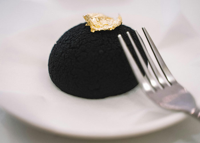 Black luxury dessert topped with edible gold leaf on a white plate with a silver fork, showcasing luxury industry rip-offs.