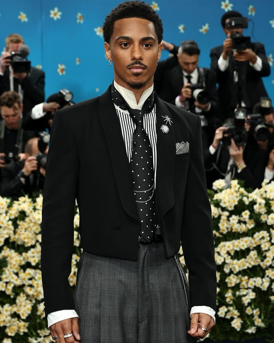 Young man in stylish black and gray outfit posing at event, surrounded by photographers and floral decorations, pop culture moment.
