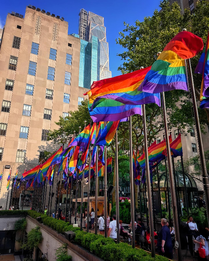 Colorful pride flags flying along a walkway in a busy NYC urban area with tall buildings and clear blue sky.