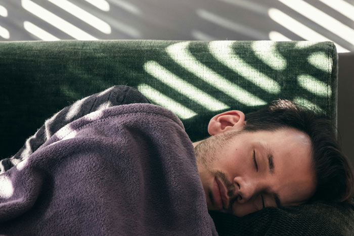 Man resting on green couch with eyes closed, wrapped in blanket, illustrating woman shutting down boys nights at their house.