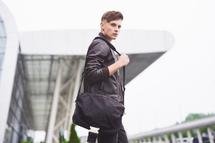 Young man carrying a bag outdoors, looking contemplative while standing near a modern building with unresolved mysteries vibes.