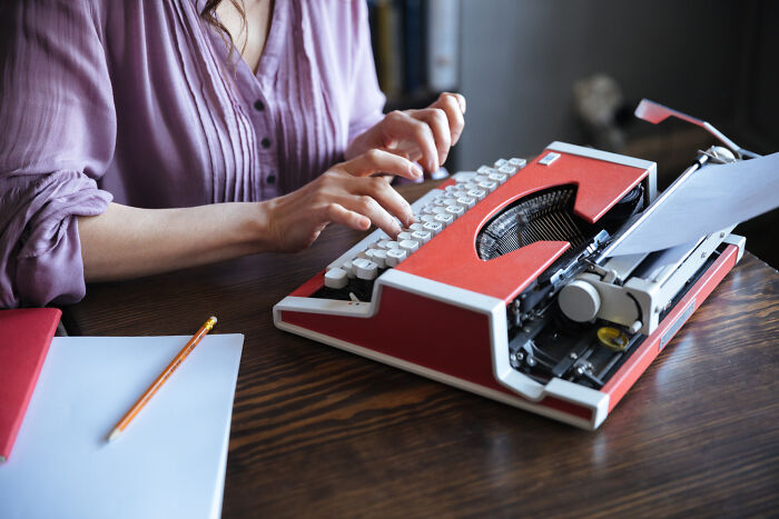 Person typing on a vintage red typewriter at a wooden desk illustrating frustrating work habits at office.