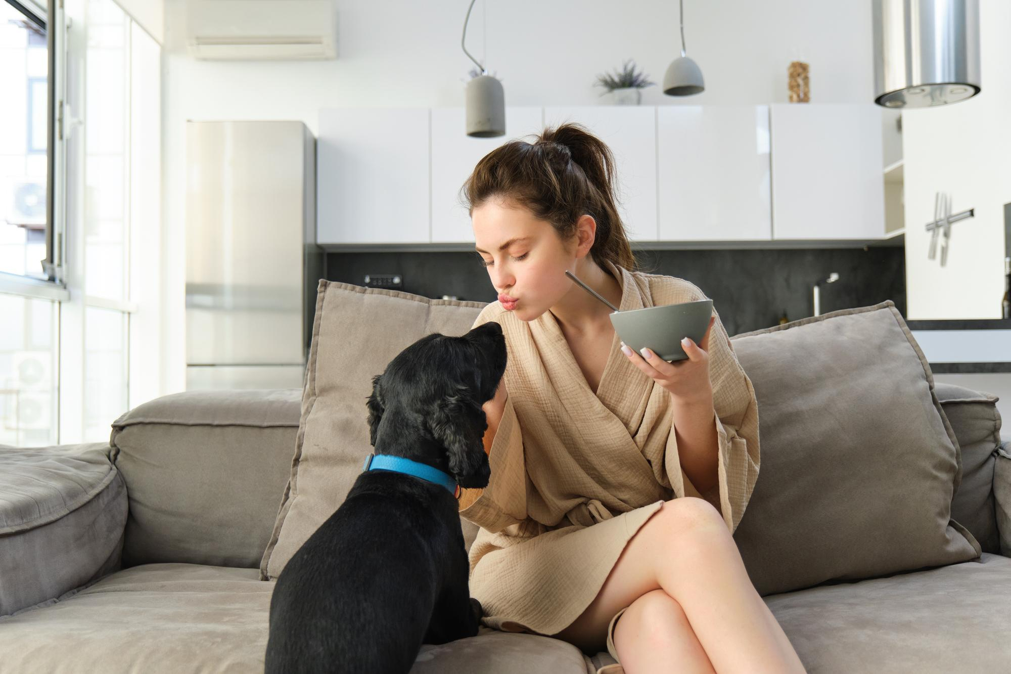 Young woman in robe sitting on sofa showing affection to black dog inside modern home dog care home drama concept