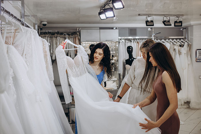 Three women examining a wedding dress during a wedding dress shopping trip signaling a painful friendship wake-up call. Three women examining a wedding dress during a wedding dress shopping trip signaling a painful friendship wake-up call.
