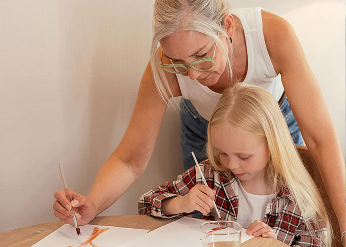 Woman with glasses helping child paint at table, capturing unhinged things their MIL just said moments.