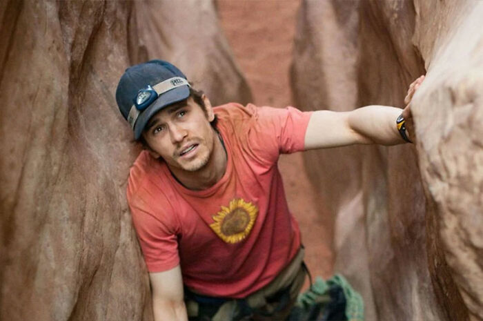 James Franco wearing a cap and red shirt, climbing between narrow rock walls in an outdoor setting. James Franco wearing a cap and red shirt, climbing between narrow rock walls in an outdoor setting.