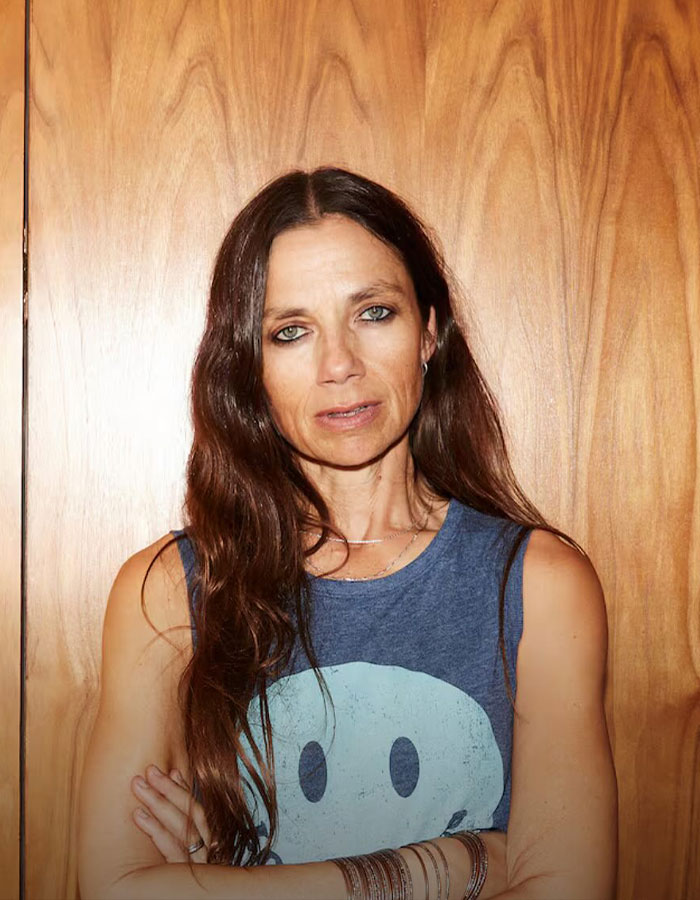 Actress Justine Bateman posing with long hair and arms crossed against a wooden background in casual clothing.