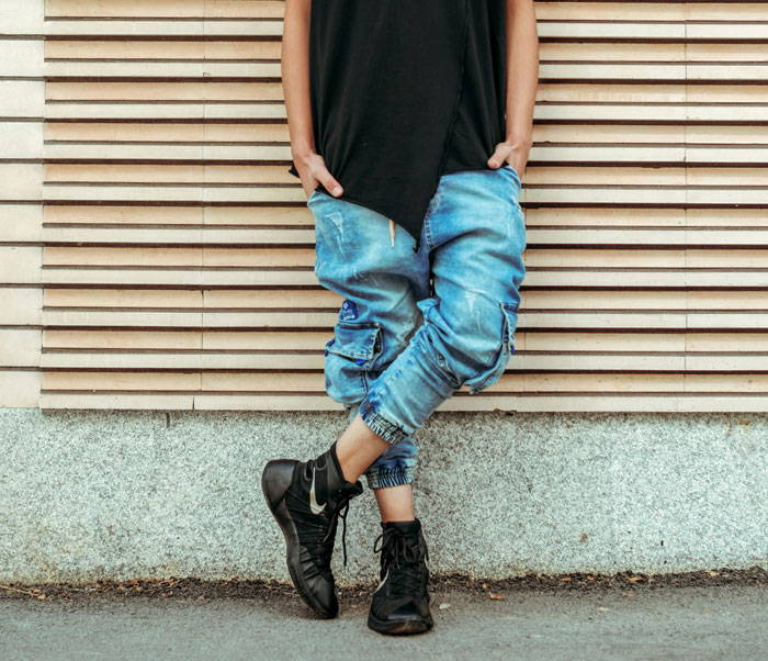 Teen wearing black shirt and blue jeans leaning against wall, representing next-level evil kids in urban setting.