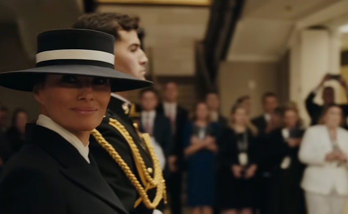 Melania Trump wearing a black hat and coat at a formal event with people in the background. Melania Trump wearing a black hat and coat at a formal event with people in the background.
