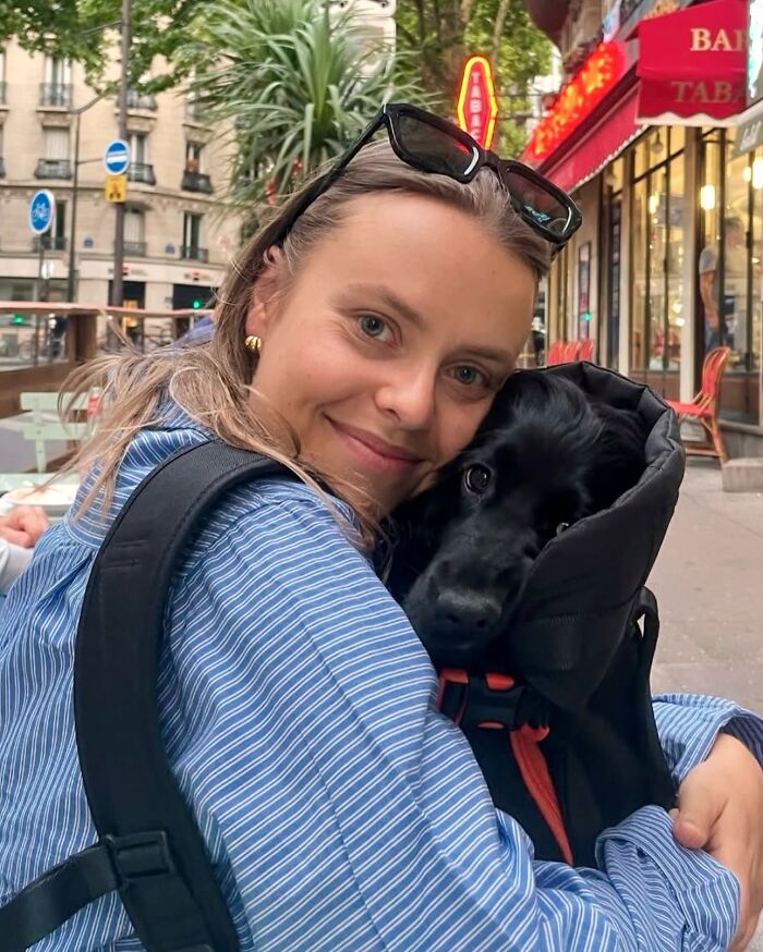 Woman hugging black dog in backpack on city street, showcasing adorable dogs in backpacks enjoying urban adventures.