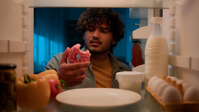 Young man looking confused holding a bitten doughnut inside a fridge, illustrating revenge stories with bitter aftertaste concept.