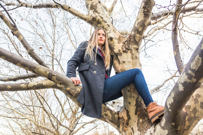 Young woman sitting on a tree branch, wearing a coat and boots, captured in an embarrassing moment seen by others.