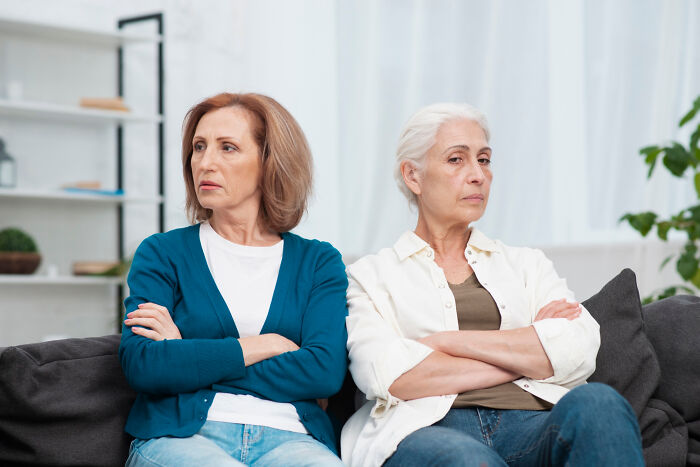 Two middle-aged women sitting apart on a couch with arms crossed, showing family drama and tension.