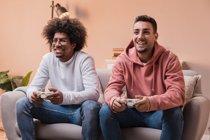 Two men playing video games on a couch, smiling and focused, illustrating men missed delivery babies scenario.