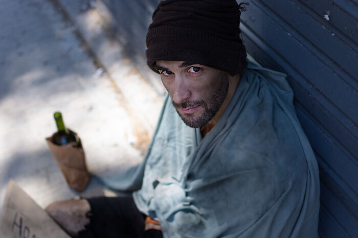 Man wrapped in a blanket sitting against a wall with a bottle nearby, one of the bizarre things seen through a neighbor's window.