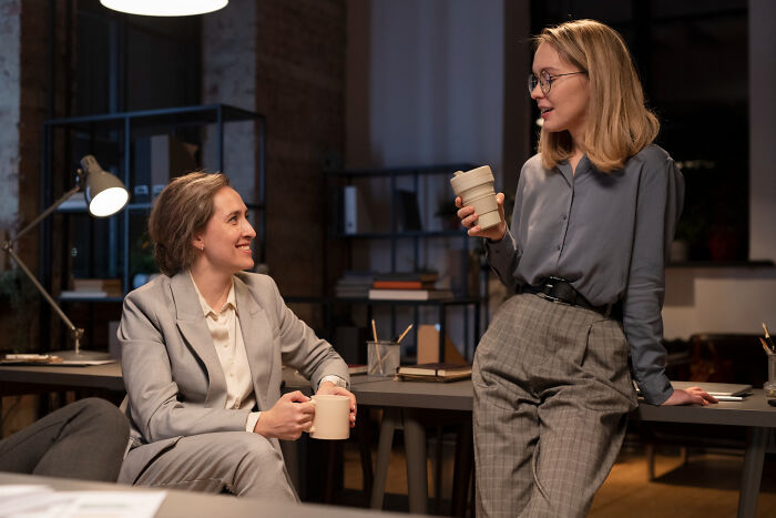 Two women chatting and drinking coffee in an office, illustrating small tricks that make being a grown-up easier.