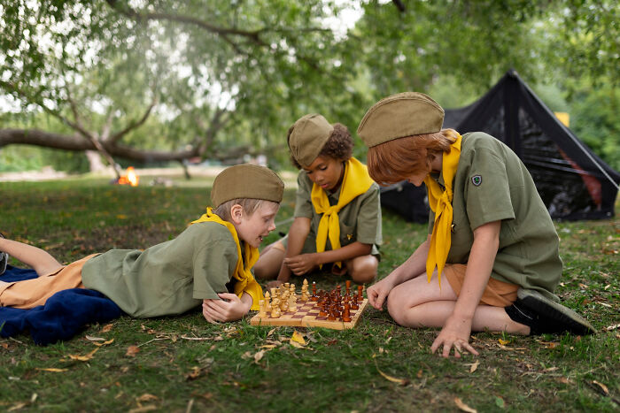 Three kids in scout uniforms playing chess outdoors, capturing embarrassing moments people were blessed to see.