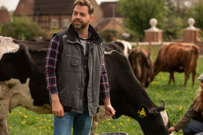Man in plaid shirt and vest holding a bucket near grazing cows, evoking unresolved mysteries people carry with them.