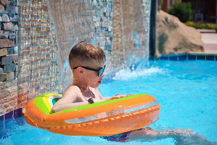 Young boy in sunglasses relaxing in a colorful inflatable ring in a pool near a waterfall feature, capturing embarrassing moments.