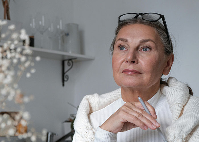 Thoughtful older woman sitting indoors, wearing glasses on her head, reflecting on unhinged things her MIL just said.