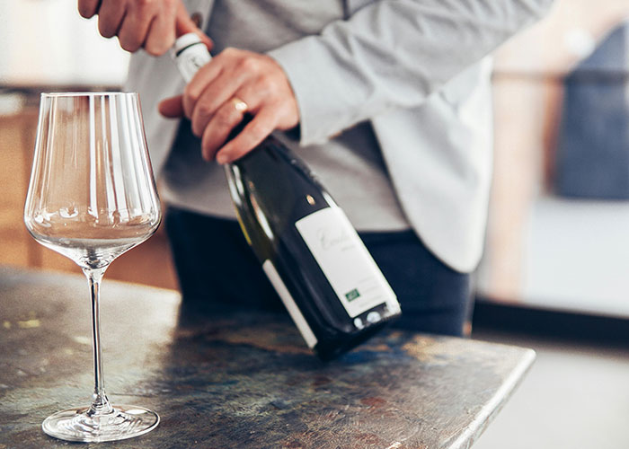 Person opening a bottle of wine next to an empty glass, illustrating luxury industries and rip-off purchases by the rich.