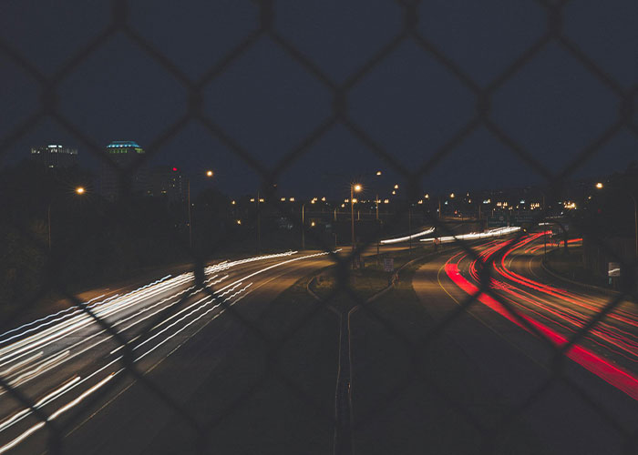 Nighttime highway light trails seen through a fence, evoking creepy and mysterious scenes in remote and isolated places.