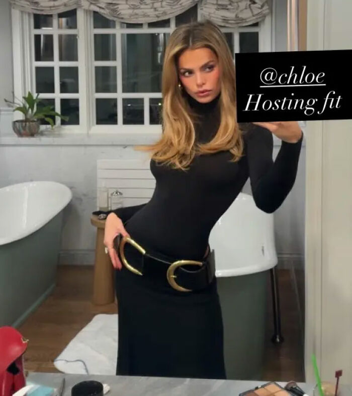 Brooks Nader posing in a black dress with a large Santa belt worn as a shirt in a bathroom mirror selfie.