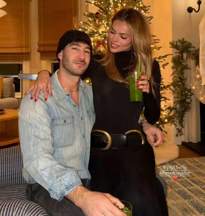 Brooks Nader wearing a Santa belt as a shirt in Aspen, posing indoors with a man during holiday season.