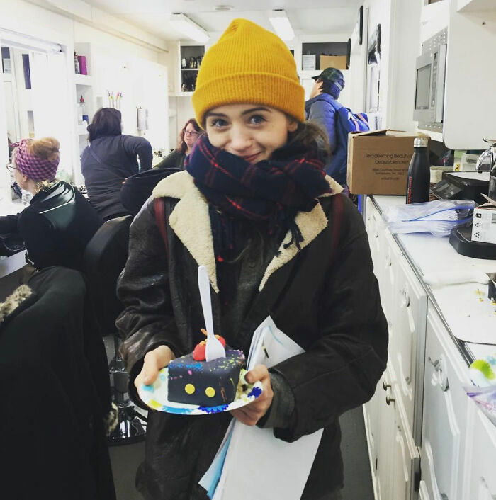Stranger Things star Natalia Dyer wearing a yellow beanie and scarf, holding a slice of cake indoors, fans concerned with appearance.