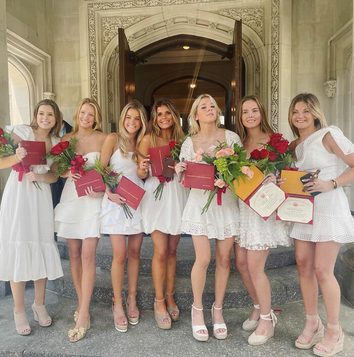 Sorority girls in white dresses holding diplomas and roses, celebrating graduation at a mansion with ornate stone architecture.