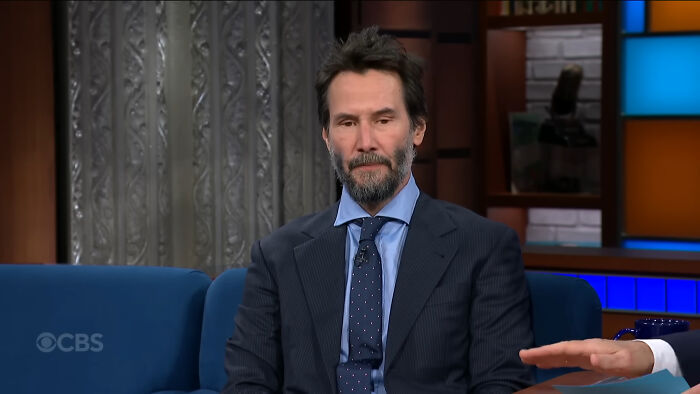 Actor Keanu Reeves in a dark suit and patterned tie during a talk show interview revealing celebrity secrets.