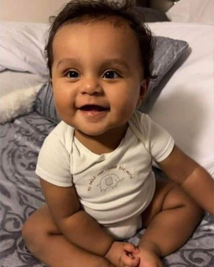 Smiling toddler sitting on a bed, wearing a white onesie, representing the accused beauty queen's case in court.