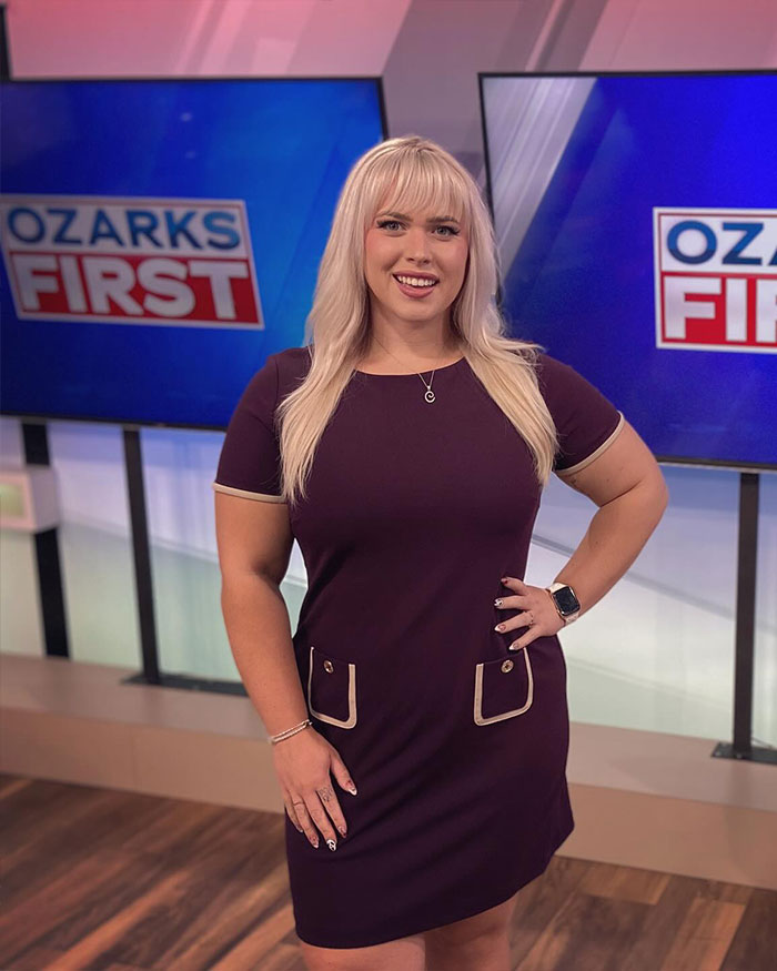 Curvy news anchor standing confidently in studio with Ozarks First screens in background, smiling at camera.
