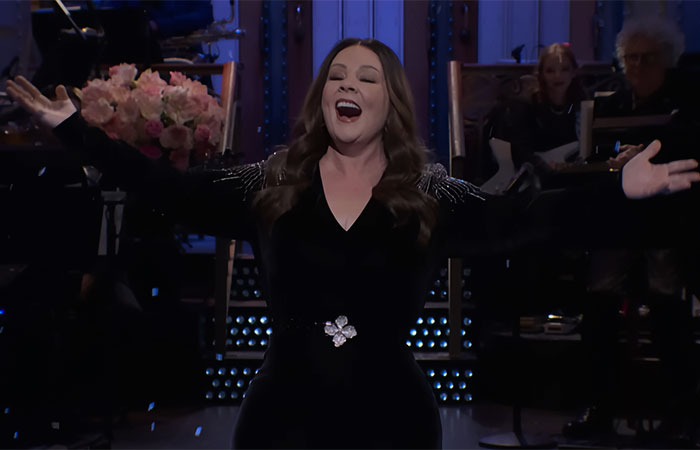 Melissa McCarthy on SNL stage wearing black dress with arms wide open, showcasing her jaw-dropping 95-pound weight loss.