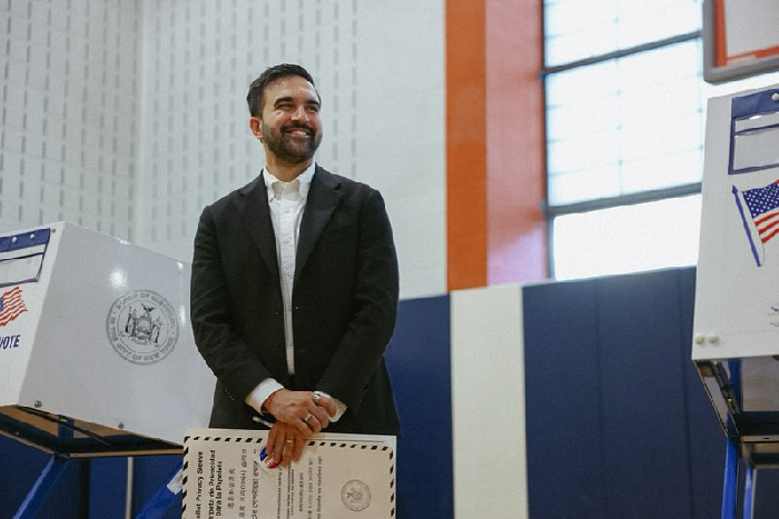 Man standing at a voting booth inside a gymnasium, smiling and holding a ballot, NYC election environment.