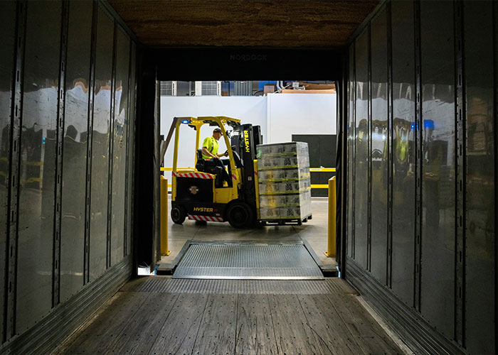 Worker operating forklift inside warehouse, handling stacked pallets during busy industrial loading process.