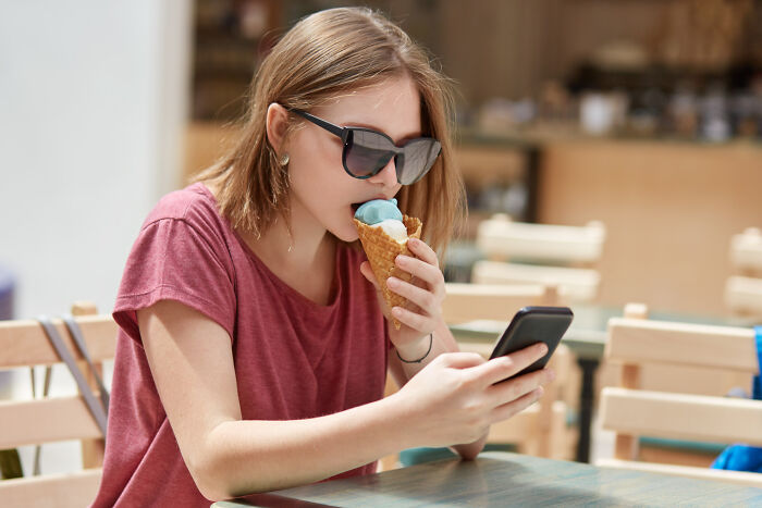 Young woman wearing sunglasses eating ice cream while looking at her phone, an embarrassing moment people were blessed to see.