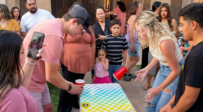 Group of people playing a popular party game outdoors with cups on a colorful game board during a social gathering.